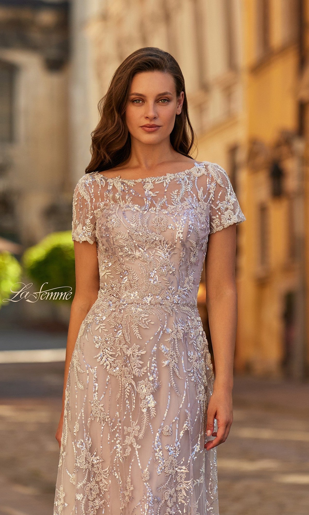 A woman in the La Femme 31838 long formal dress with short sleeves and beaded embroidery stands outdoors on a sunlit, building-lined street.
